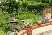 A small informal suburban country garden with a pond, shrubs and perennials with climbers on a boundary fence, view over low patio wall 