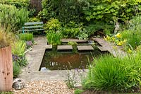 A small garden pond with stepping stones in a suburban country garden, surrounded with stone paving, a garden seat, and informal mixed planting of perennials and shrubs