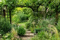 Stepping stones lead under the arch of a wooden pergola with bird box. Borders of perennials and grasses in an informal cottage-garden style