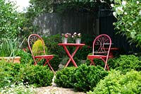 Red, metal table and chairs in garden, with low informal border of Japanese Box plants.