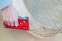 Woman using staple gun to attach chicken wire to wooden sieve. 