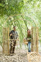 Man and a woman holding bamboo craft objects under a bamboo arch in a clearing of living Phyllostachys - Bamboo