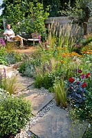 Man sitting on a chair in garden, view over path to flower bed. Planting includes: Stipa tenuissima, Dahlia 'HS Romeo', Erigeron karvinskianus, Buxus sempervirens, Salvia nemorosa 'Caradonna', Briza media, Lavandula angustifolia 'Richard Gray' - Lavender, Calamagrostis brachytricha and Penstemon 'Raven' 