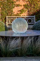 Aqualens water feature designed by Allison Armour with large clipped balls of Buxus sempervirens, Stipa tenuissima grass and Traschelospermum jasminoides on brick wall, on a frosty morning.