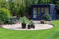 View over neat lawn and patio to a hand-built garden room. Agaves and a young Cordyline in black ceramic pots on the circular patio, Bamboo screen behind summerhouse.