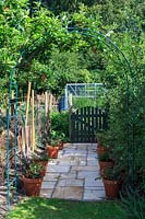 Paved pathway to kitchen garden, flanked by several compost heaps and leaf mould piles, metal arch and wooden gate