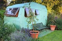 Super Sprite caravan which serves as a home office, with paved seating area with Malus 'Red Jade' - Crabapple - in terracotta pots and a bed of including Lavandula x intermedia 'Grosso' - Lavender and Caryopteris clandonensis