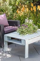 Wooden pallet table with integral sunken  planter with summer planting on slate patio in summer accompanied by outdoor chairs.