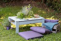 Table made out of a wooden pallet, with central planter planted with mixed herbs in summer country garden with cushions. 