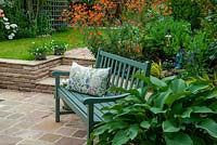 Paved patio with garden bench with floral pattern cushion and Hosta in container against a raised bed of orange Geum and other perennials. Steps lead from patio to a small lawn - Open Gardens Day, Drinkstone, Suffolk