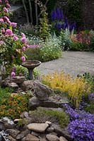 View over rocks and birdbath to flowering borders in small paved courtyard garden. 