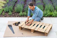 Woman using nails and hammer to fix pieces of narrow wood strips to fill gaps in pallet. 