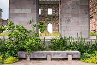 Oak bench in front of castle ruin surrounded by Acanthus mollis Latifolius Group 'Rue Ledan', Alchemilla mollis, Jasminum officinale 'Devon Cream' and Cercis canadensis f. alba 'Royal White'