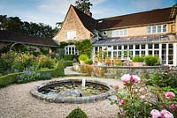 Circular pond with water spouts surrounded by box-edged beds of roses, salvias and hardy geraniums in June