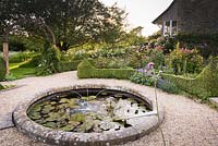 Circular pond with water spouts surrounded by box-edged beds of roses, salvias and hardy geraniums in June
