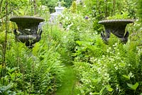 Decorative urns in borders packed with plants either side of grass path. 