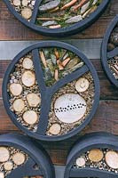 Circular insect hotels attached to a wooden wall and filled with bamboo canes, branches, stones and moss.