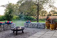 Edge of paved patio with a lit firepit on a pebble surface with gabion benches, view of garden beyond 