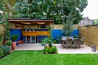 Patio, table and chairs and outdoor room with covered barbecue. Surrounded by a contemporary wooden trellis fence and blue painted wall.