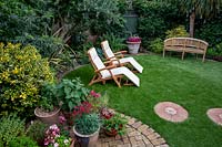 Small garden with circular artificial lawn, looking towards sun loungers and bench, area enclosed with mixed border. In foreground, small patio with container plants