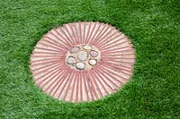 Circular terracotta path step with pebbles at centre set in artificial lawn