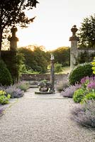 View up wide path at sunset with pools of Nepeta - Catmint - spilling from the double border and a framework of architectural fragments 