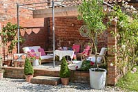 Raised seating area with metal pergola and shade cover against a brick wall, with lounge furniture and pots