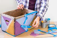 Woman removing masking tape to reveal wooden box painted in a multicoloured geometric pattern
