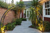 View through circular metal arch to paved area by house, beds with Cordyline and lighting 
