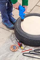 Woman using an electric drill to make pilot holes through plywood disk into car tyre
