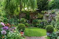 Circular lawn and rear patio enclosed in borders of roses, hardy geraniums, foxgloves, ferns and peonies, in the shade of a weeping birch. Pots of clipped small-leaved hollies, Ilex crenata 'Kinme'.