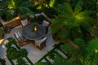 A bird's-eye view of an enclosed courtyard dining area, with the fronds of Dicksonia antarctica - Tree Fern - filtering the light from uplighters 