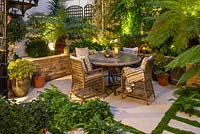 A walled courtyard with outdoor dining area lit at night, illuminating Dicksonia antartica - Tree Fern, Hydrangea and Hosta in raised beds