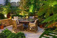 A walled courtyard with outdoor dining area lit at night, illuminating Dicksonia antarctica - Tree Fern, Hydrangea and Hosta in raised beds