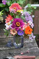 Posy of Zinnia, Hardy Geranium, Hydrangea Serrata, Schizostylis, Pelargonium and Tropaoleum in antique glass jar on rustic table in garden