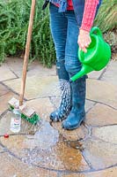 Woman watering patio with a diluted mix of white vinegar and water to clean up the paving