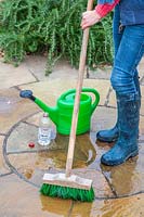 Woman using a brush to scrub a patio after having applied a mixture of white vinegar and water to clean the paving