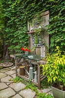 Moulded clear glass sink in old wooden vanity with red Impatiens - Balsam flowers in metal vase and Ampelopsis - Virgin vines growing on wall in backyard garden
