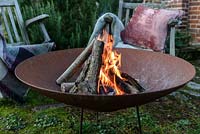 A lit fire bowl made from rusted corten steel, wooden garden chairs with wool rugs and a cushion 