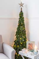 Space saving Christmas Tree made with pine - Abies and conifer - Juniperus foliage attached to a willow obelisk and decorated with star decorations and lights in a modern living room setting with chair and side table