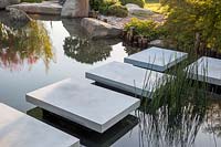 Floating stone stepping stones over a pond with Equisetum hyemale, beyond large stone boulders. A Japanese Reflection Garden