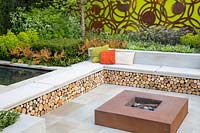 Large sawn Yorkstone stone patio with corten steel fire pit and a bench with colourful cushions and log storage underneath.The Sunken Retreat. RHS Malvern Spring Festival, 2016. Design: Ann Walker for Graduate Gardeners
