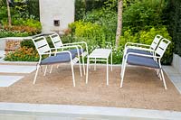 White metal framed table and chairs with blue cushions on crushed stone gravel seating area. The Sunken Retreat. RHS Malvern Spring Festival, 2016. Design: Ann Walker for Graduate Gardeners
