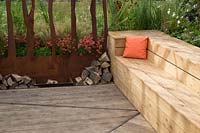 A timber framed wooden bench seating area with orange cushion, by firepit with logs and a corten steel screen divider. Prospect and Refuge garden, RHS Tatton Park Flower Show, 2017. Designer: Anca Panait