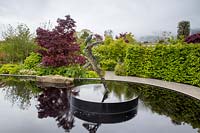 Reflective black pond water feature with sculpture 'Zephyr' by Simon Gudgeon on a misty May spring day - mixed planting - garden hedge and Acer palmatum 'Bloodgood' Garden of Quiet Contemplation. RHS Malvern Spring Festival 2019 - Designer Peter Dowle - Leaf Creative