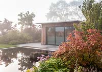 Wooden garden office studio in front of a pool and paved stone patio area on a misty May morning - planted with Acer palmatum 'Shishigashira' in foreground and shrubs. Garden of Quiet Contemplation. RHS Malvern Spring Festival 2019  - Designer Peter Dowle - Leaf Creative 
