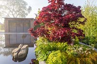 Office studio in front of reflective black pond water feature on a misty May spring day - mixed planted border with grasses ferns and green foliage plants - Acer palmatum 'Bloodgood' Garden of Quiet Contemplation garden. RHS Malvern Spring Festival 2019  - Designer Peter Dowle - Leaf Creative 