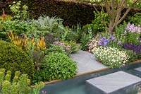 A Sustainable garden based around herbaceous planting - grey stone garden path with stepping stone over a rill water feature surrounded by mixed flower borders - Sicillian chamomile -Iris 'Jane Phillips' - Centaurea montana, Cynara cardunculus, Euphorbia, Fagus sylvatica f. purpurea - Copper Beech hedge. The Morgan Stanley Garden. RHS Chelsea Flower Show 2019 - Designer: Chris Beardshaw - Sponsor: Morgan Stanley 