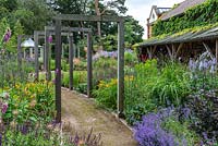 A contemporary lower garden with a path leading beneath a pergola made up of eight standalone oak frames that frame views over the borders of Aconites, Daylilies, Dahlias, Geum, Sea hollies, Catmint, Campanulas, Salvias and Verbenas.