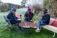 Women wrapped in warm clothes, roasting marshmallows over a fire in a Corten Steel fire pit on a frosty winter day. 